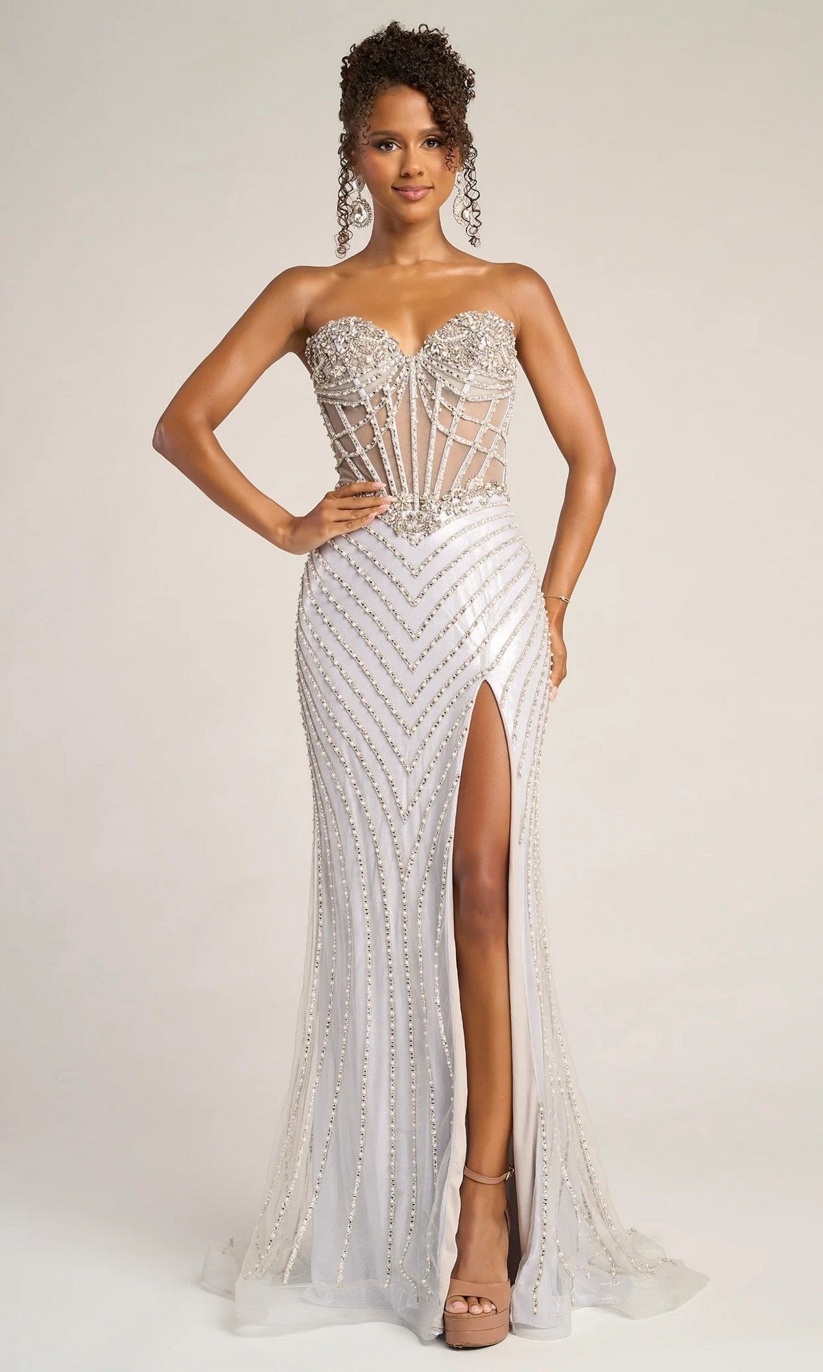 A woman poses confidently with one hand on her hip, wearing the Portia and Scarlett XO057 long formal dress in silver, featuring a strapless design, sheer corset bodice, beadwork, and a thigh-high slit against a plain background.